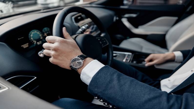 Close up top view of man's watch in blue suit keeping hand on the steering wheel while driving a luxury car.